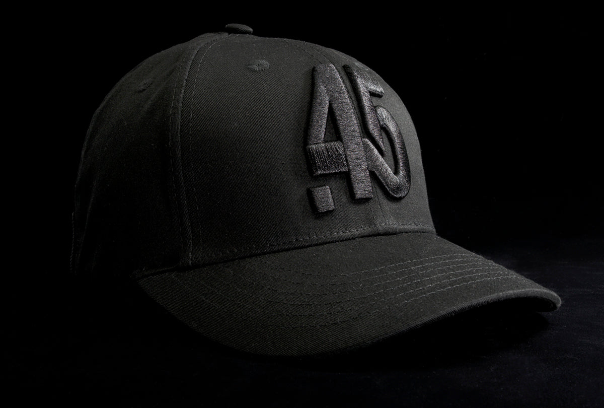 A Fourfive 45 Knight Racer hat with flat brim in all black stretch fit material and a revolver cylinder embroidery detail in the front with the hat facing 45 degrees to the right in dramatic lighting with black background.  Built to fit normal and big heads.