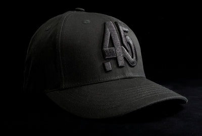 A Fourfive 45 Knight Racer hat with flat brim in all black stretch fit material and a revolver cylinder embroidery detail in the front with the hat facing 45 degrees to the right in dramatic lighting with black background.  Built to fit normal and big heads.