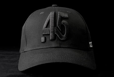 A Fourfive 45 Knight Racer hat with curved brim in all black stretch fit material and 45 logo embroidery detail in the front with the hat facing straight in dramatic lighting with black background.  Built to fit normal and big heads.