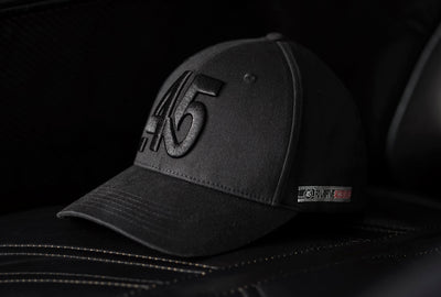 A Fourfive 45 Knight Racer hat with curved brim in black stretch fit material and an embroidered 45 logo in the front facing 45 degrees to the right sitting on a black leather sofa with dramatic lighting.  Built to fit normal and big heads.