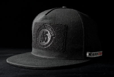 A fourfice 45 Customizer hat shadow edition with flat brim black cotton front with black velcro logo detail for customising and black mesh back with snapback facing 45 degrees left in dramatic lighting with black background.  Built to fit normal and big heads.