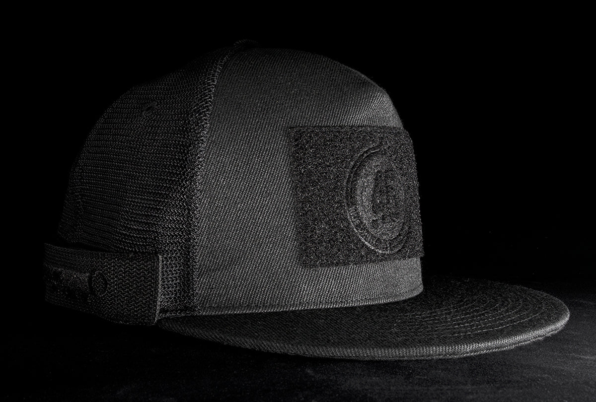 A fourfice 45 Customizer hat shadow edition with flat brim black cotton front with black velcro logo detail for customising and black mesh back with snapback facing 45 degrees right in dramatic lighting with black background.  Built to fit normal and big heads.