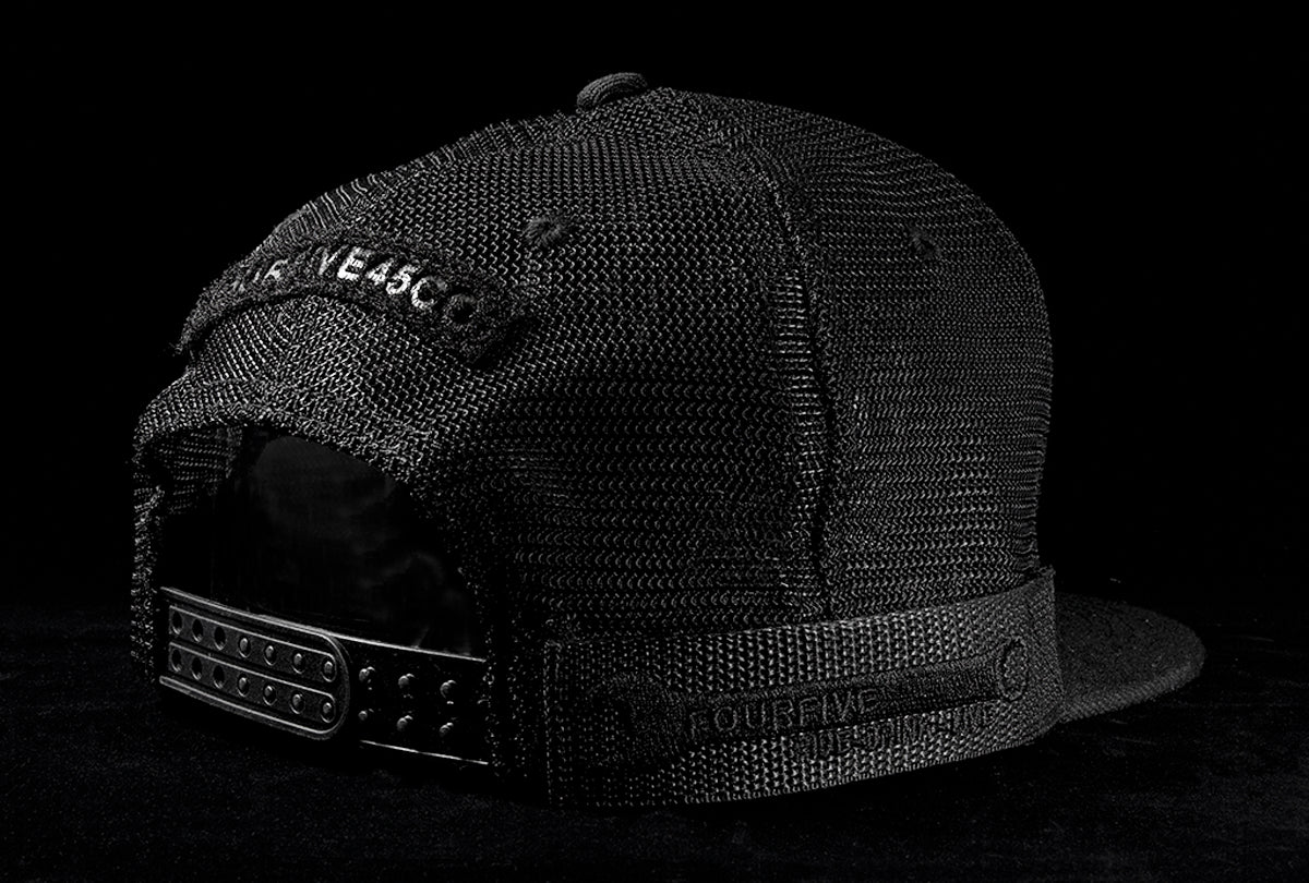 A Fourfive 45 Customizer hat shadow edition with flat brim black cotton front and black velcro logo detail for cuctomising and black mesh back with snapback with its 45 degree back view showing a velcro detail and the hat holster mount with its embroidery details in dramatic lighting with black background.  Built to fit normal and big heads.