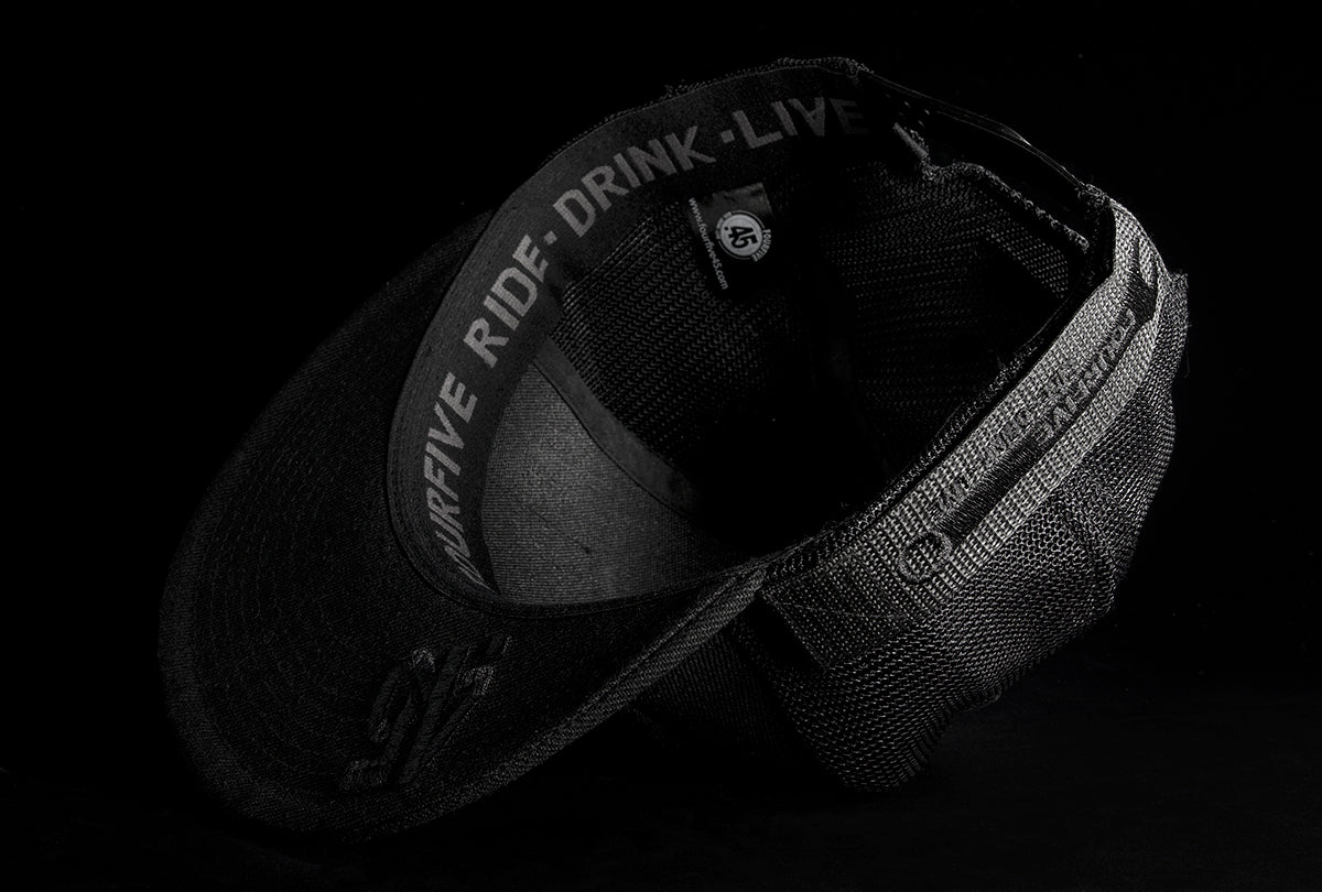 A Fourfive 45 Customizer hat shadow edition with flat brim black cotton front and black velcro logo detail for customising and black mesh back  flipped over showing the 45 logo embroidery detail on the bottom of the brim and the ride drink live embroidery detail around the sweat band on the inside of the hat.  Built to fit normal and big heads.