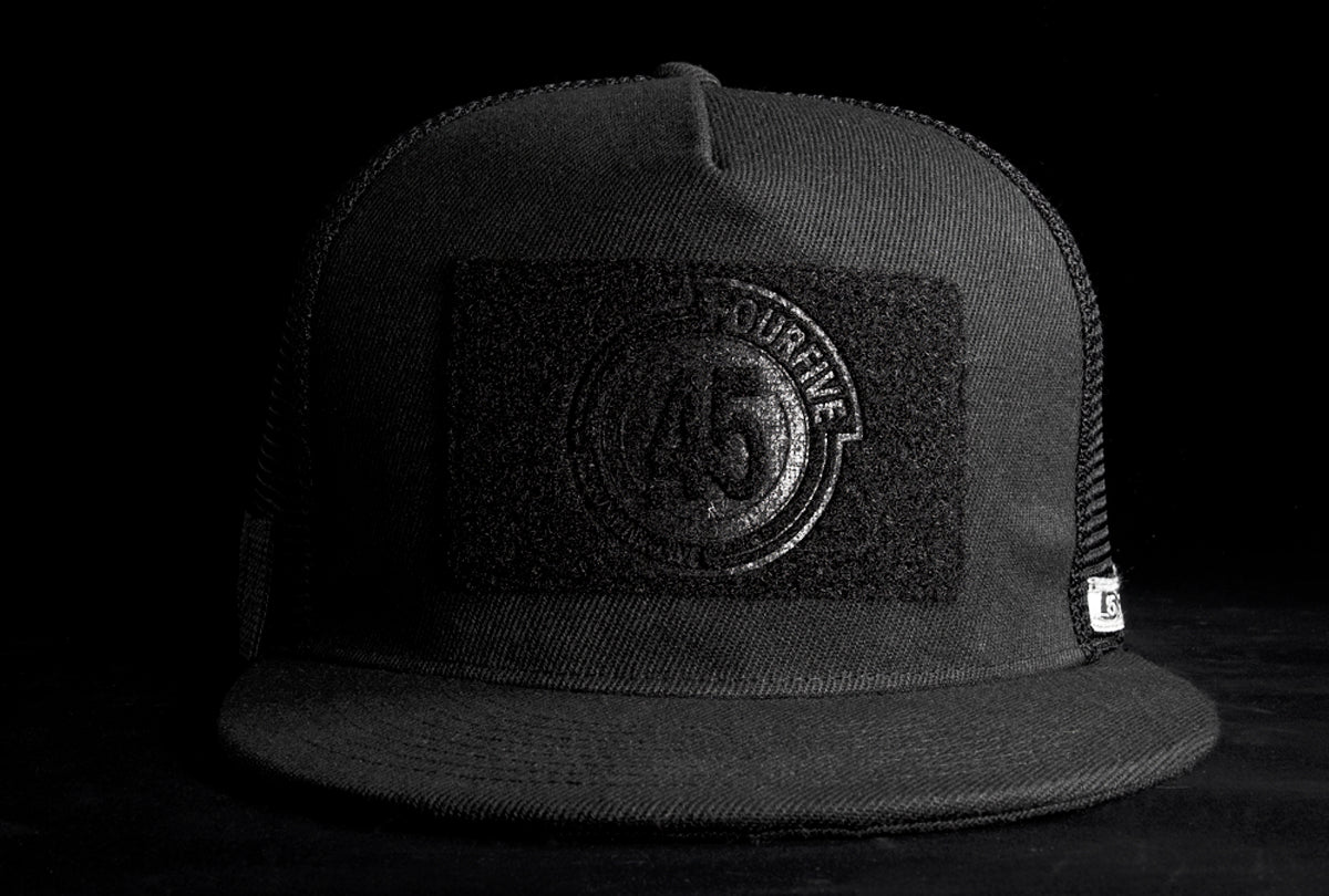 A Fourfive 45 Customizer hat shadow edition with flat brim black cotton front with black velcro logo detail for customising with your own velcro patches facing straight in a black background with dramatic lighting.  Built to fit normal and big heads.