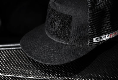 A Fourfive 45 Customizer hat shadow edition with flat brim black cotton front and black velcro logo detail for customising with your own velcro patches and black mesh back and snapback sitting on a carbon fibre armchair.   Built to fit normal and big heads.