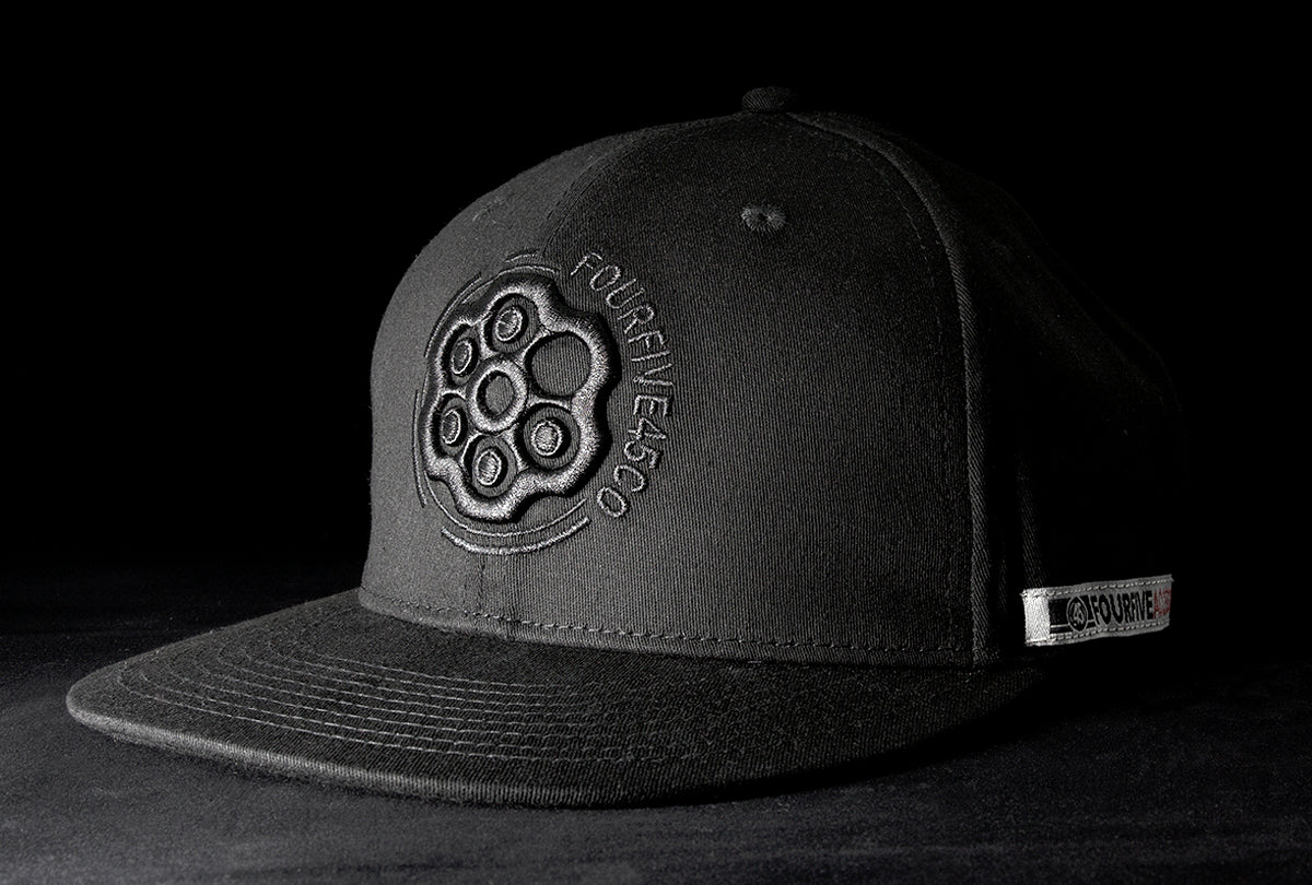 A Fourfive 45 Revolver hat black edition with flat brim in all black stretch fit material and a revolver cylinder embroidery detail in the front with the hat facing 45 degrees to the right in dramatic lighting with black background.  Built to fit normal and big heads.