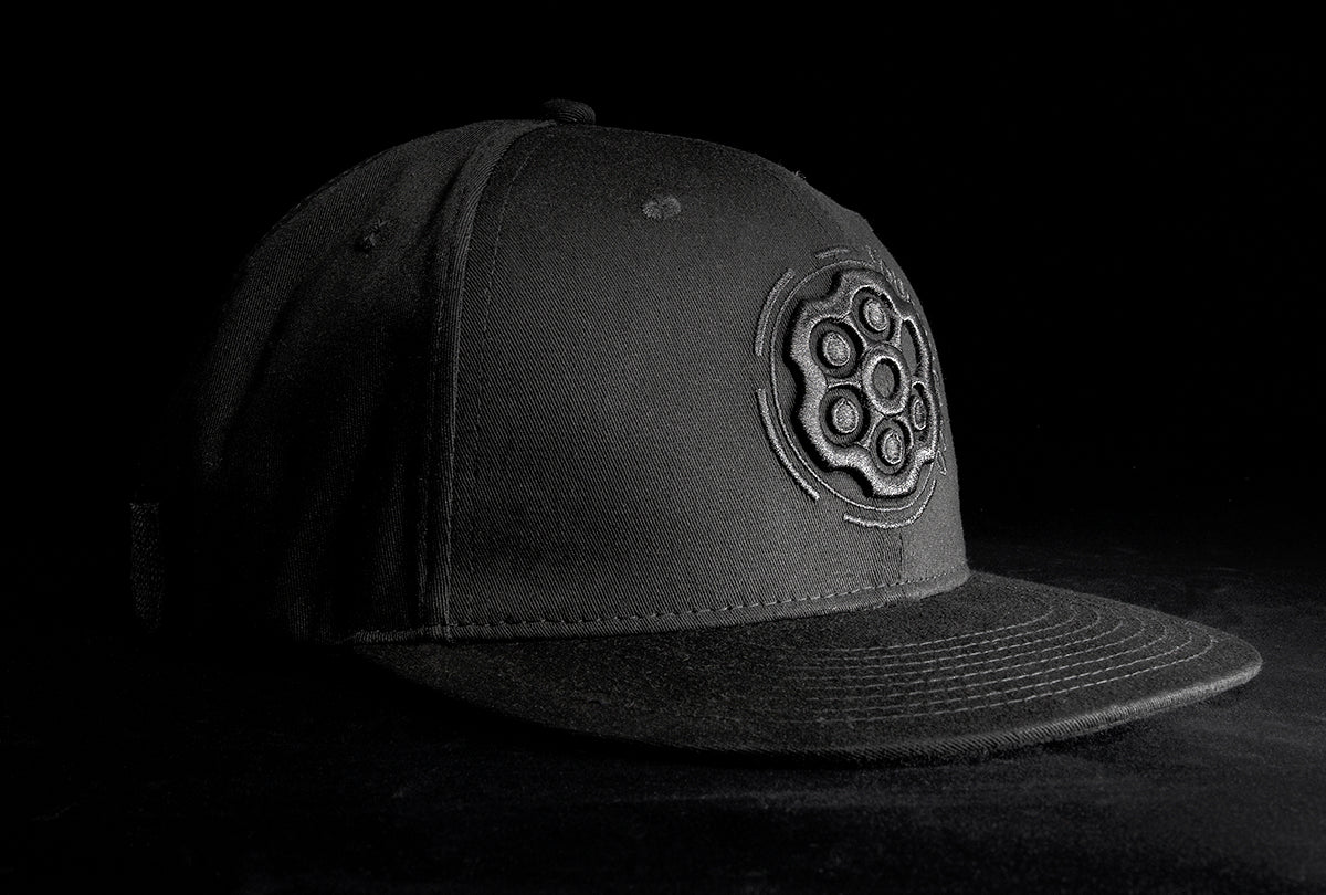 A Fourfive 45 Revolver hat black edition with flat brim in all black stretch fit material and a revolver cylinder embroidery detail in the front with the hat facing 45 degrees to the left in dramatic lighting with black background.  Built to fit normal and big heads.