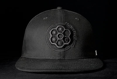A Fourfive 45 Revolver hat black edition with flat brim in all black stretch fit material and a revolver cylinder embroidery detail in the front with the hat facing straight in dramatic lighting with black background.  Built to fit normal and big heads.