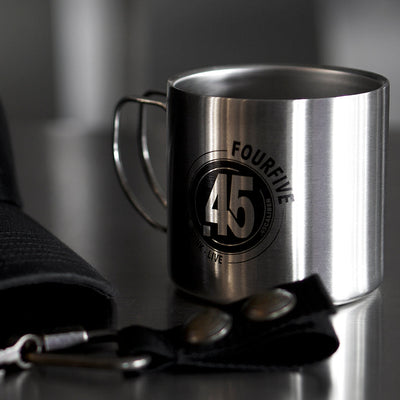 A Fourfive 45 ACP Coffee mug in double walled stainless steel construction with a Fourfive 45 logo printed on its body and a our of focus hat holster in front both sitting on a stainless steel table with dramatic lighting.