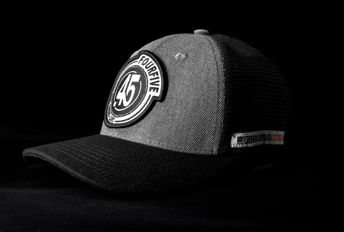 A Fourfive 45 Flat Tracker hat with a Curved brim in a grey cotton material front with a Fourfive logo embroidery detail in the front and black mesh back with a snapback strap and the hat facing 45 degrees to the left in dramatic lighting with black background.  Built to fit normal and big heads.