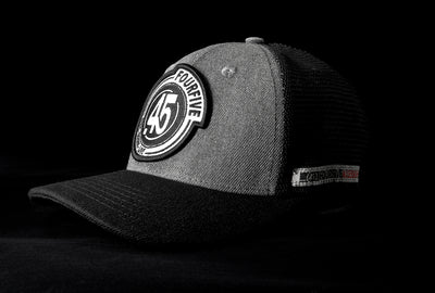A Fourfive 45 Flat Tracker hat with a Curved brim in a grey cotton material front with a Fourfive logo embroidery detail in the front and black mesh back with a snapback strap and the hat facing 45 degrees to the left in dramatic lighting with black background.  Built to fit normal and big heads.