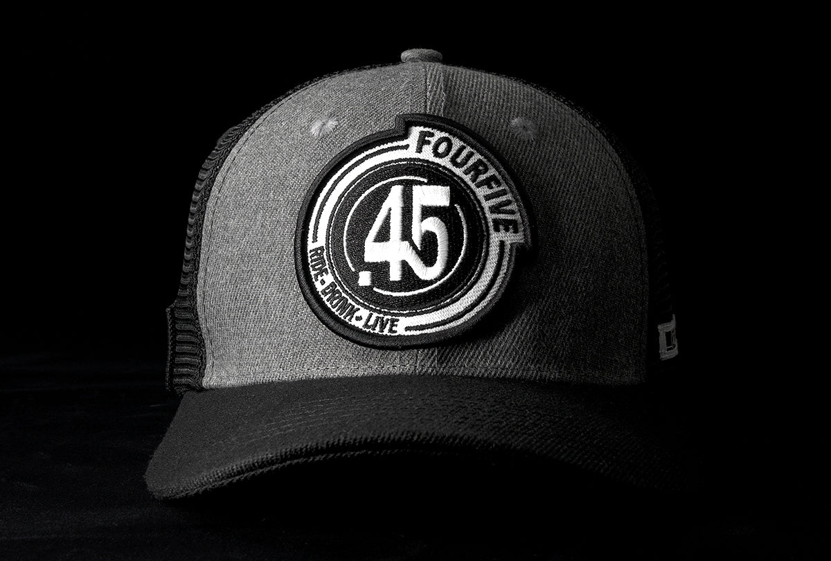 A Fourfive 45 Flat Tracker hat with a curved brim in a grey cotton material front with a Fourfive logo embroidery detail in the front and black mesh back with a snapback strap and the hat facing straight in dramatic lighting with black background.  Built to fit normal and big heads.