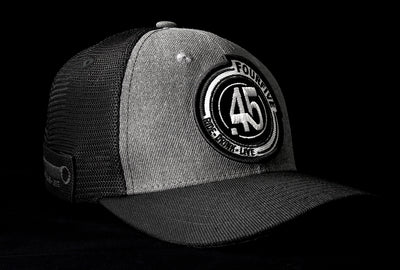 A Fourfive 45 Flat Tracker hat with a curved brim in a grey cotton  material front with a Fourfibe logo embroidery detail in the front and black mesh back with a snapback strap and the hat facing 45 degrees to the right in dramatic lighting with black background.  Built to fit normal and big heads.