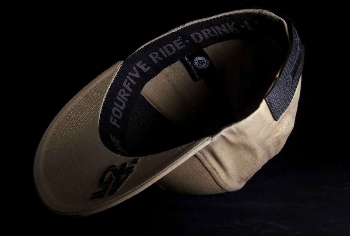 A Fourfive 45 Revolver hat sand edition with flat brim in a khaki coloured stretch fit material flipped over showing the 45 logo embroidery detail on the bottom of the brim and the ride drink live embroidery detail around the sweat band on the inside of the hat.  Built to fit normal and big heads.