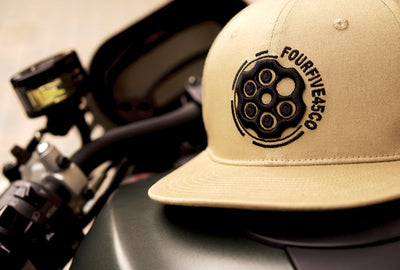A Fourfive 45 Revolver hat sand edition with flat brim in a khaki coloured stretch fit material with a revolver cylinder embroidery detail on the front the hat facing slightly to the left while sitting on the fuel tank of a Ducati Monster motorcycle in dusk lighting.  Built to fit normal and big heads.