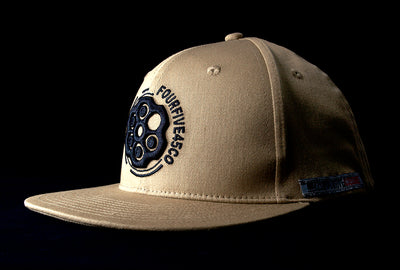 A Fourfive 45 Revolver hat sand edition with flat brim in a khaki coloured stretch fit material and a revolver cylinder embroidery detail in the front with the hat facing 45 degrees to the left in dramatic lighting with black background.  Built to fit normal and big heads.
