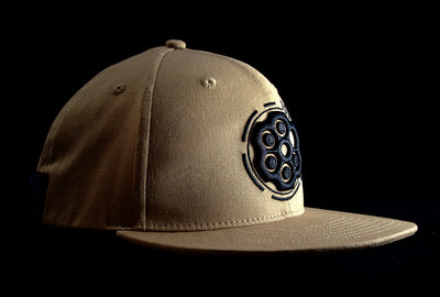 A Fourfive 45 Revolver hat sand edition with flat brim in a khaki coloured stretch fit material and a revolver cylinder embroidery detail in the front with the hat facing 45 degrees to the right in dramatic lighting with black background.  Built to fit normal and big heads.