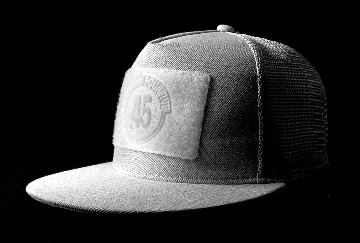 A fourfice 45 Customizer hat snow edition with flat brim light grey cotton front with white velcro logo detail for customising and light grey mesh back with snapback facing 45 degrees left in dramatic lighting with black background.  Built to fit normal and big heads.