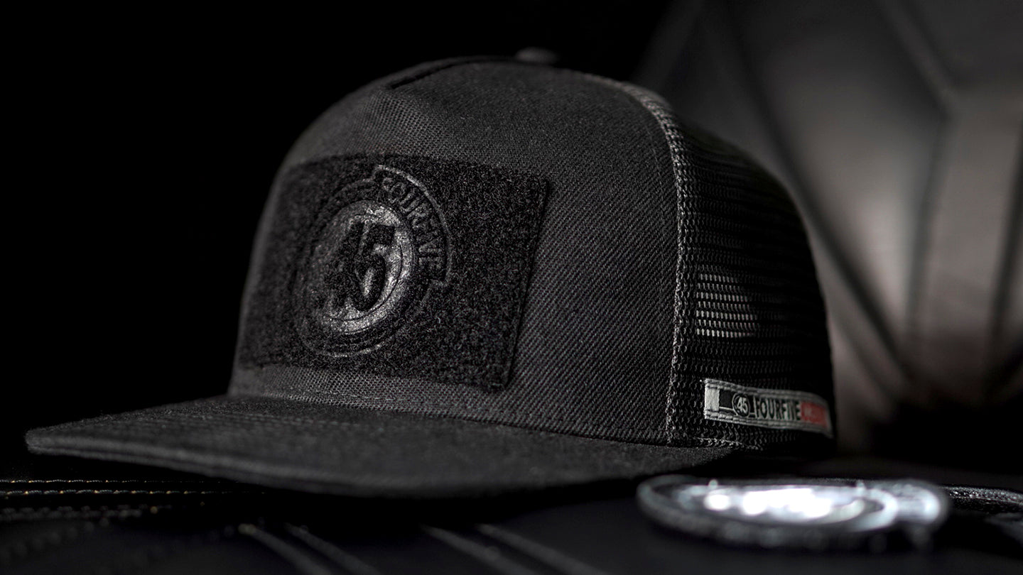 Fourfive 45, all black flat brim snap back customizer hat with velcro elements on front and back sitting on a black leather armchair.
