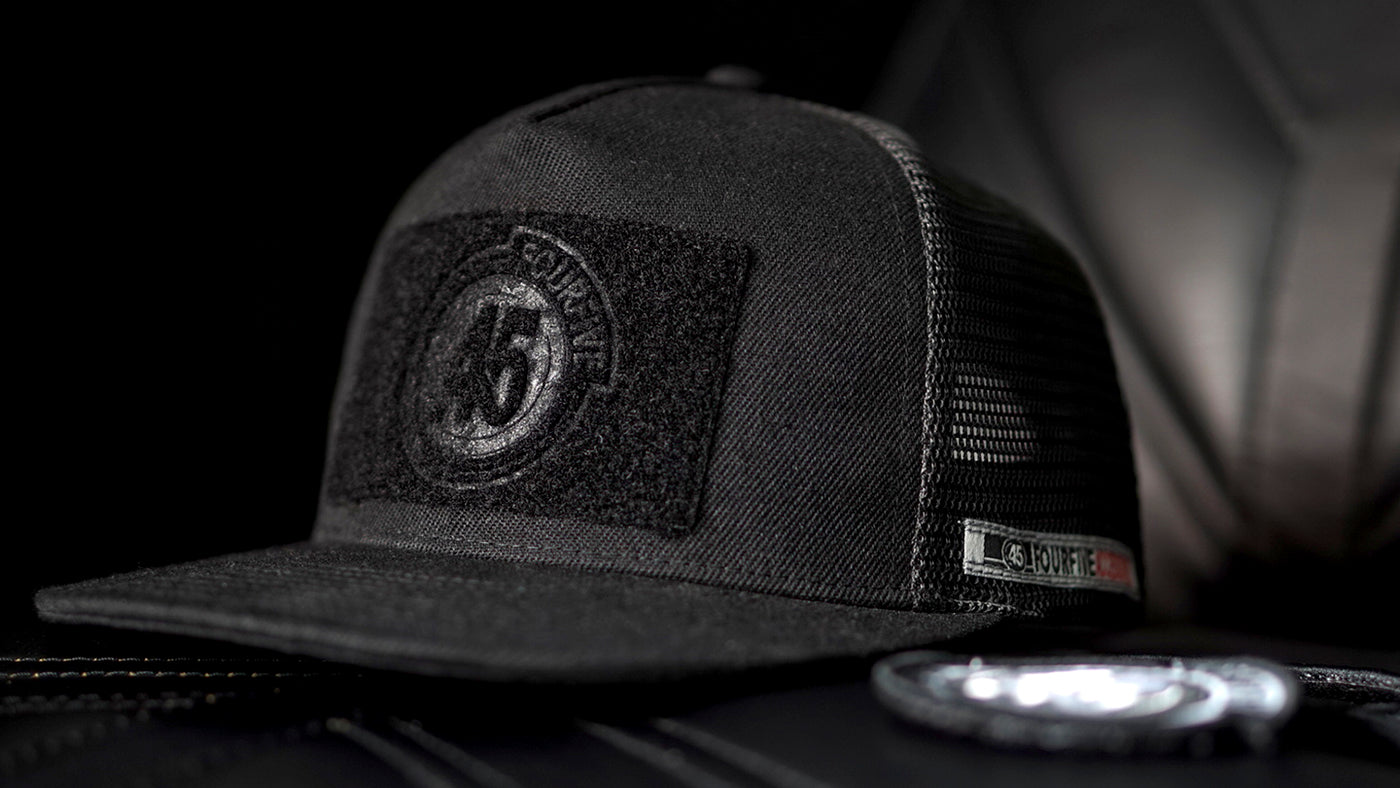 Fourfive 45, all black flat brim snap back customizer hat with velcro elements on front and back sitting on a black leather armchair.