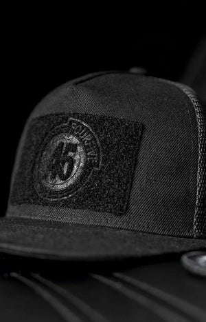 Fourfive 45, all black flat brim snap back customizer hat with velcro elements on front and back sitting on a black leather armchair.