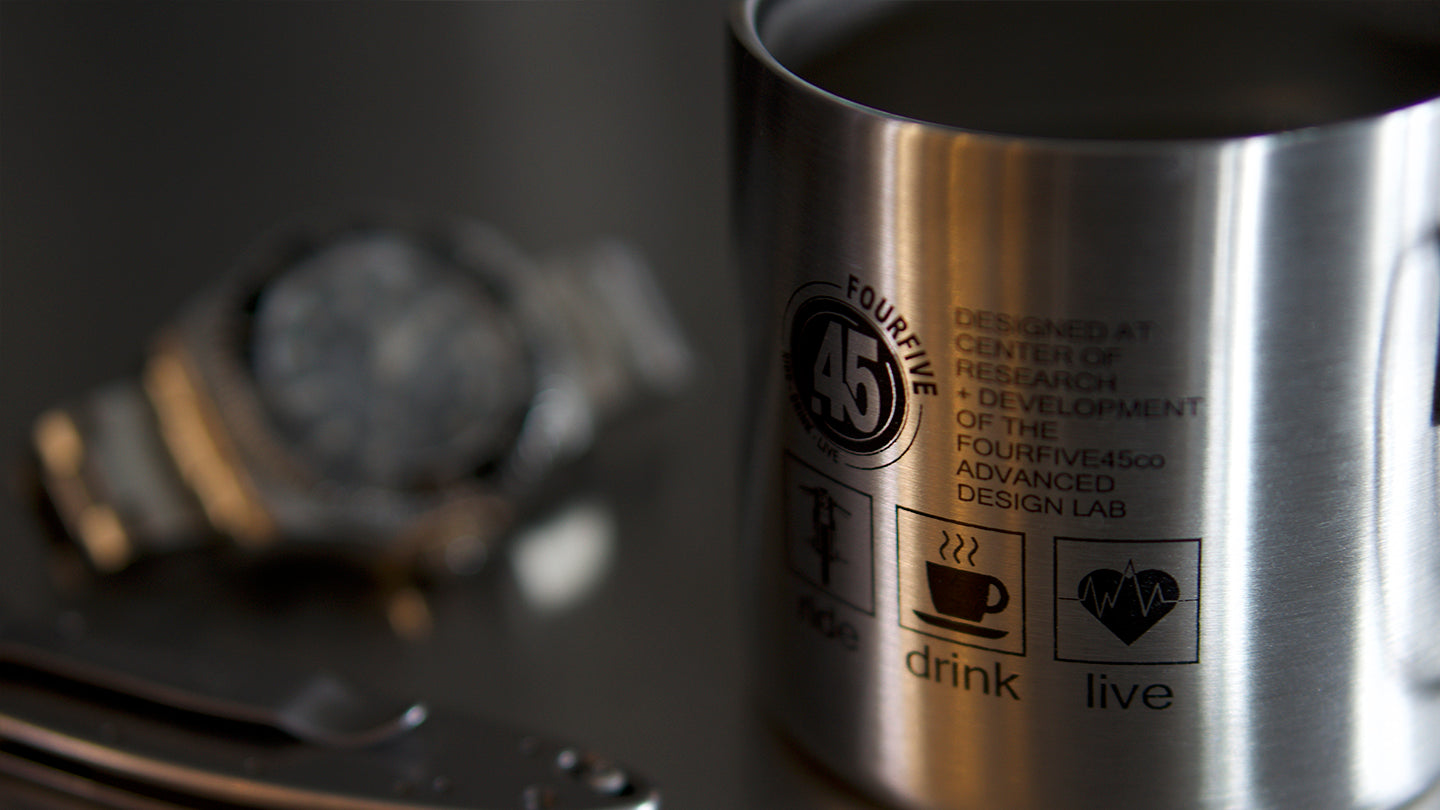 Stainless steel double walled coffee mug by Fourfive 45, with printed graphics on front and back in dusk lighting.  A blurred rolex submariner in back ground and tactical blade in the front.