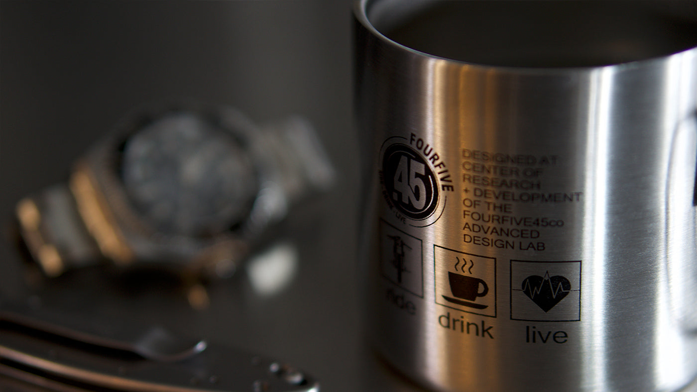 Stainless steel double walled coffee mug by Fourfive 45, with printed graphics on front and back in dusk lighting.  A blurred rolex submariner in back ground and tactical blade in the front.