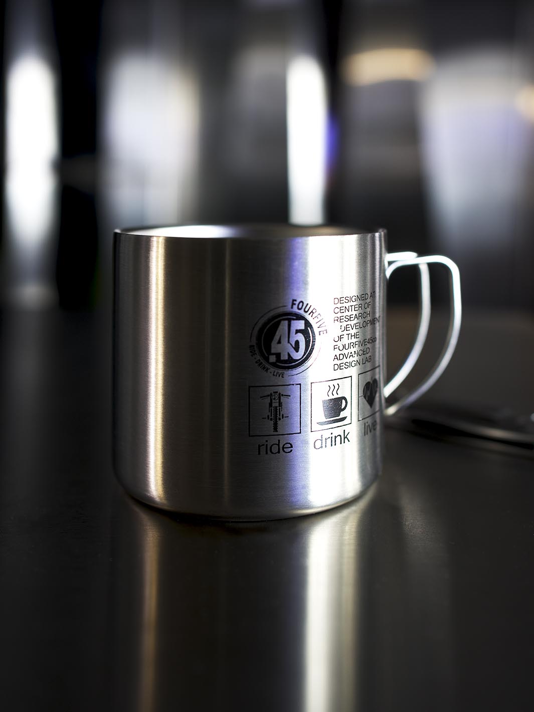 Stainless steel double walled coffee mug by Fourfive 45 with printed graphics on front and back sitting on steel table with dramatic lighting and a blurred back ground.