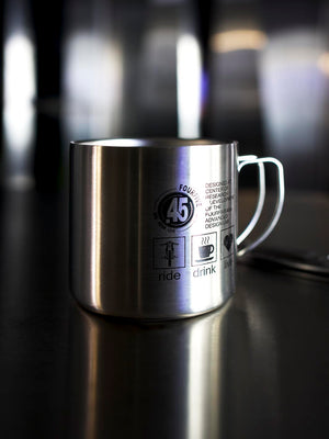 Stainless steel double walled coffee mug by Fourfive 45 with printed graphics on front and back sitting on steel table with dramatic lighting and a blurred back ground.