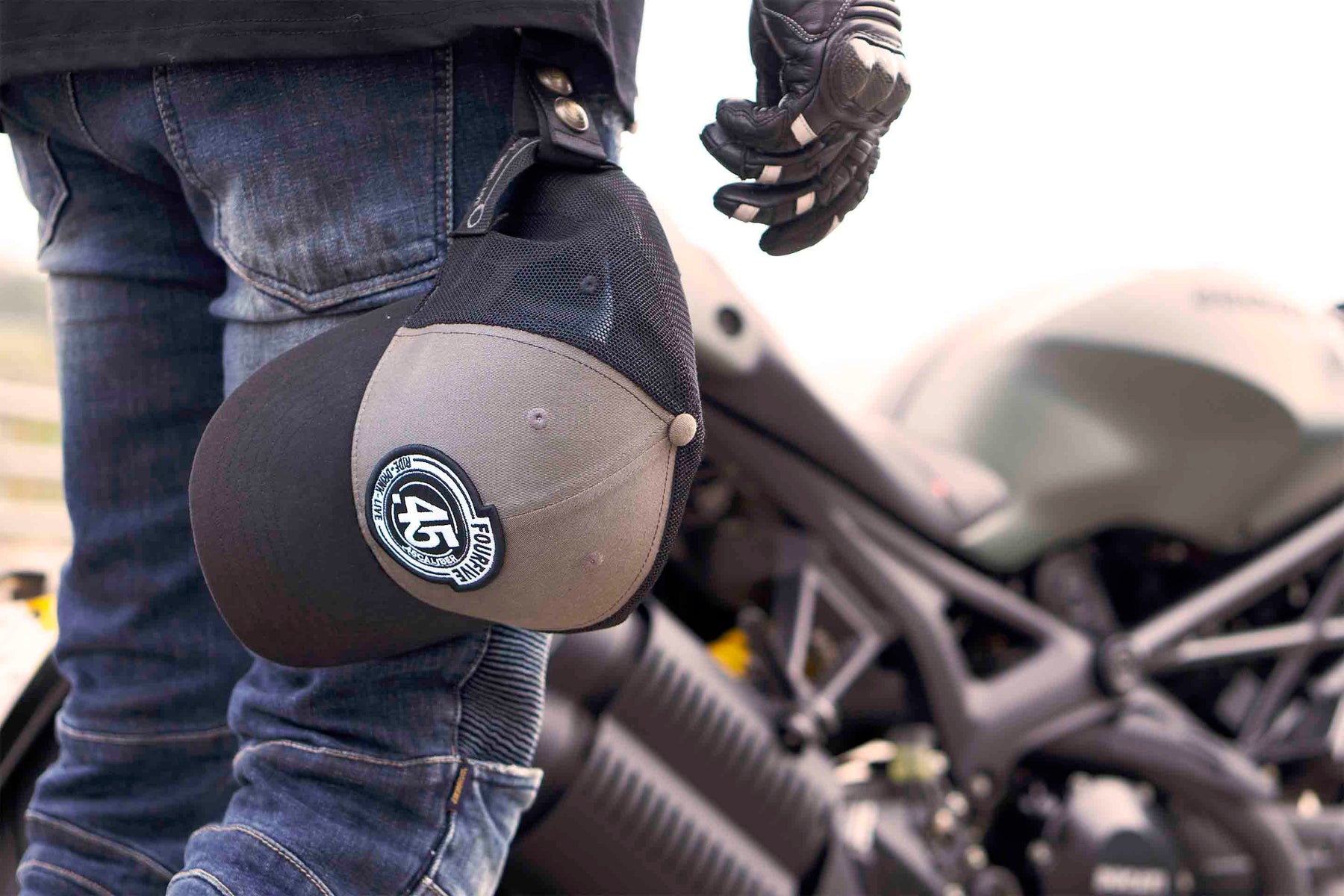 A Fourfive 45 grey and black Flat tracker trucker snap back hat holstered to a Fourfive Hat holster as rider walks towards a Ducati Monster in a dusk setting..