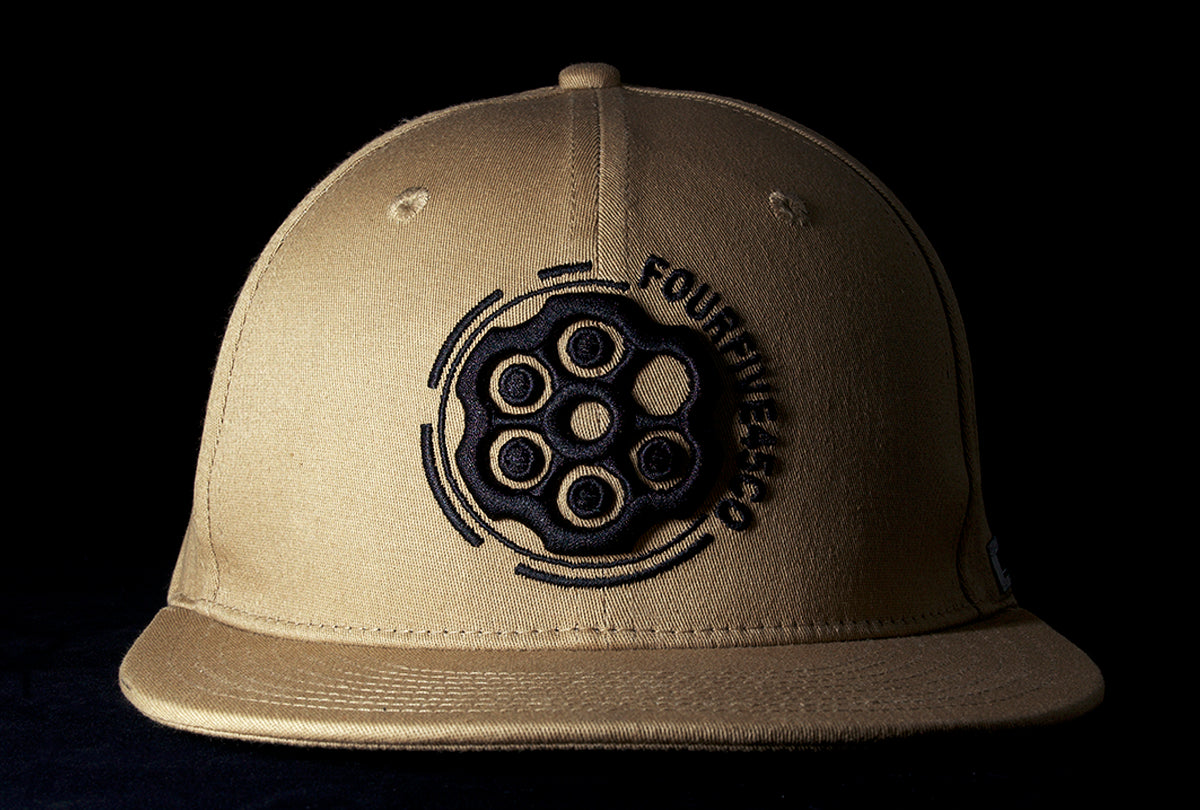 A Fourfive 45 Revolver hat sand edition with flat brim in a khaki coloured stretch fit material and a revolver cylinder embroidery detail in the front with the hat facing straight in dramatic lighting with black background.  Built to fit normal and big heads.
