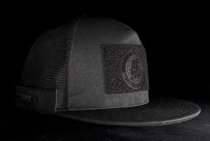 A fourfice 45 Customizer hat shadow edition with flat brim black cotton front with black velcro logo detail for customising and black mesh back with snapback facing 45 degrees right in dramatic lighting with black background.  Built to fit normal and big heads.