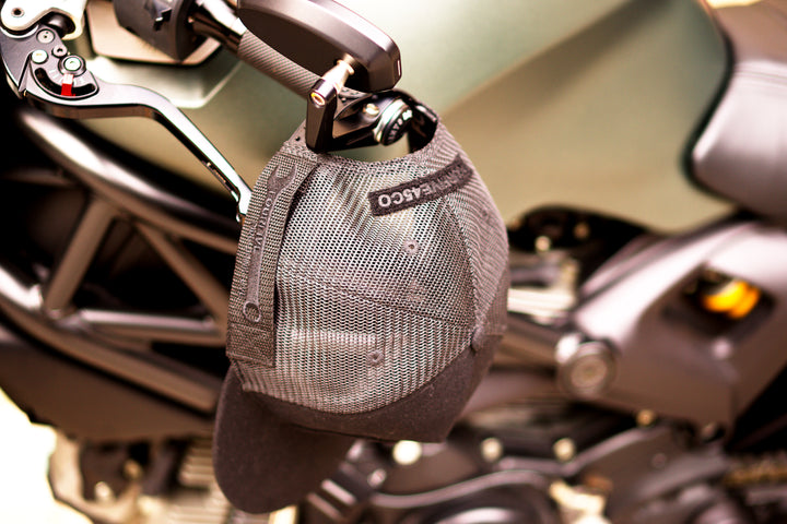 A Fourfive 45 Customizer hat shadow edition with flat brim black cotton front and black velcro logo detail for customising and black mesh back hanging on the handle bars of a green ducati monsters in dusk lighting.  Built to fit normal and big heads.